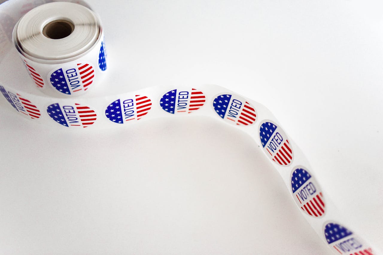 who-we-are Close-up of a roll of 'I Voted' stickers featuring the American flag design on a white background.