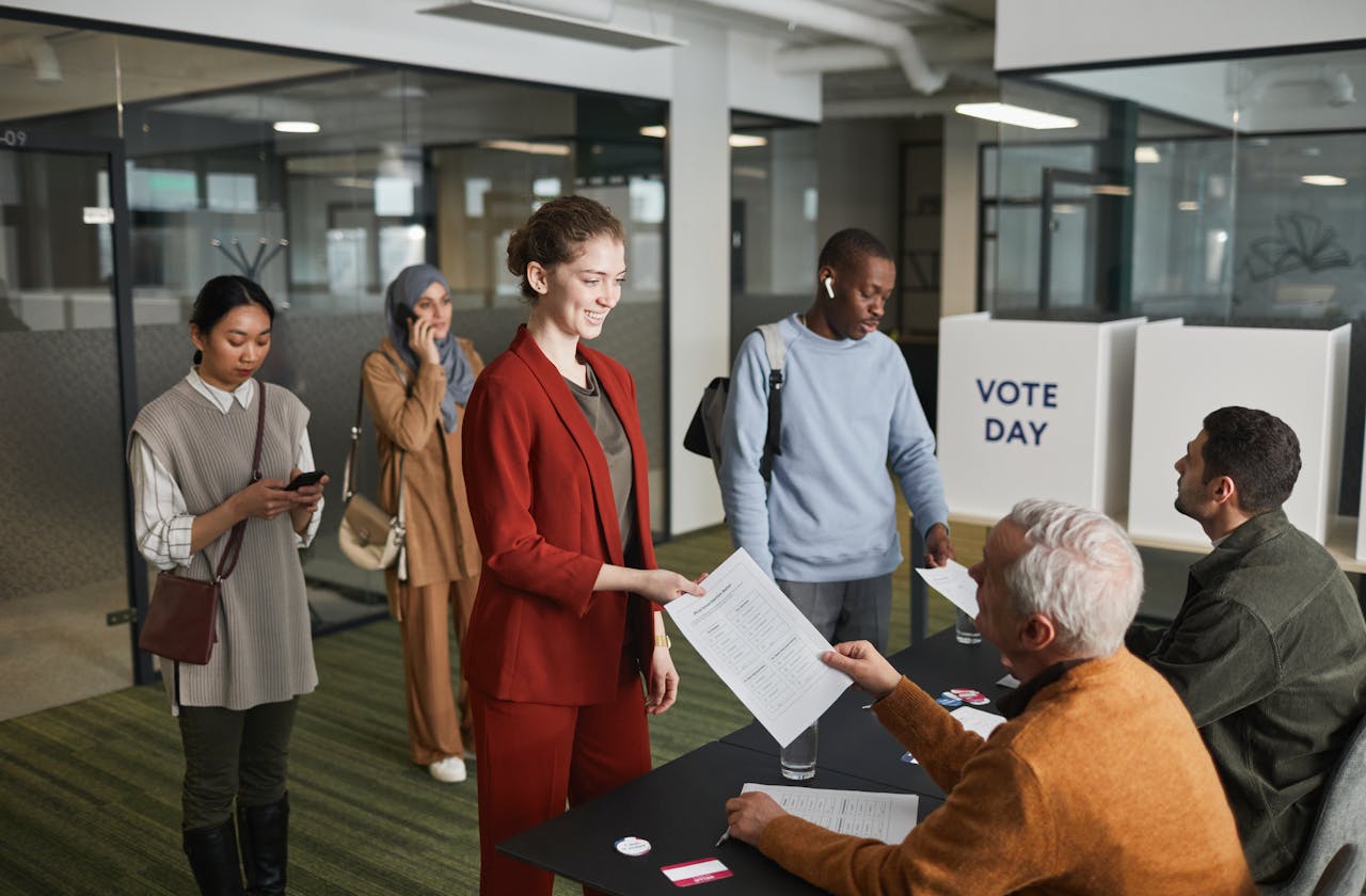 services-01 A diverse group of adults participating in an indoor voting process, engaging with election officials.